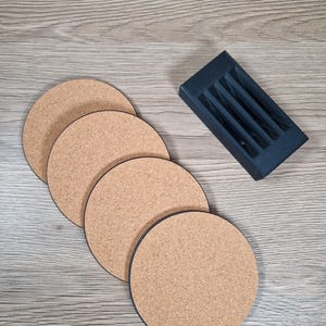 May include: Four round cork coasters with black edges are arranged on a wooden surface. A black holder with slots for pens or pencils sits to the side. The coasters are approximately 4 inches in diameter.