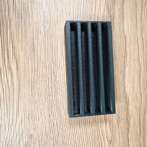 May include: A black rectangular object with five vertical slots. The object has a textured surface and is set against a light brown wooden background. The slots appear to be evenly spaced and run the length of the object.