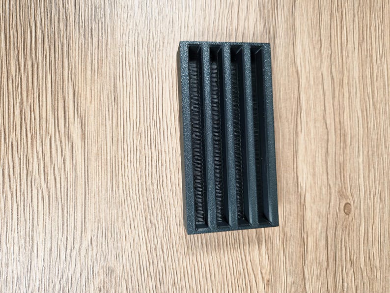 May include: A black rectangular object with five vertical slots. The object has a textured surface and is set against a light brown wooden background. The slots appear to be evenly spaced and run the length of the object.
