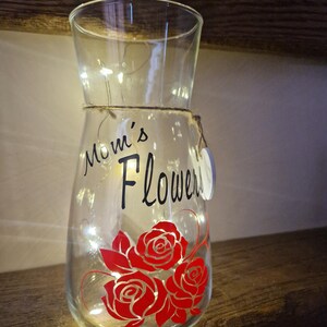 May include: A clear glass vase with a string of white lights inside. The vase has a black vinyl decal that says "Mom's Flowers" and a red rose design.