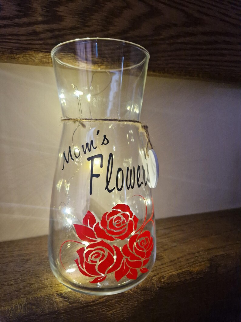 May include: A clear glass vase with a string of white lights inside. The vase has a black vinyl decal that says "Mom's Flowers" and a red rose design.