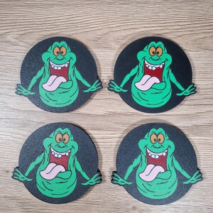 May include: Four round coasters featuring a green cartoon ghost character with a pink tongue and a wide grin. The character is set against a black background. The coasters are arranged on a wooden surface.