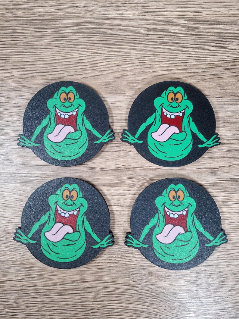 May include: Four round coasters featuring a green cartoon ghost character with a pink tongue and a wide grin. The character is set against a black background. The coasters are arranged on a wooden surface.