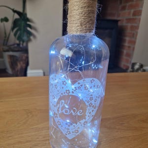 May include: A clear glass bottle with a heart design and the word "love" etched on it. The bottle is wrapped with twine and has white fairy lights inside.