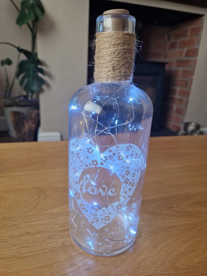 May include: A clear glass bottle with a heart design and the word "love" etched on it. The bottle is wrapped with twine and has white fairy lights inside.