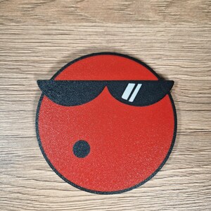 May include: A red, circular coaster with a black border and a graphic of a face wearing black sunglasses. The face has a small black dot on the bottom left side.