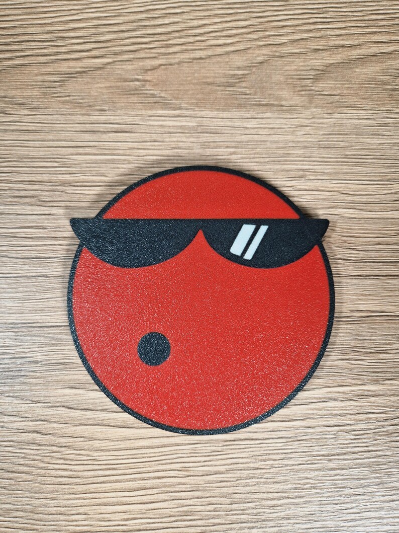 May include: A red, circular coaster with a black border and a graphic of a face wearing black sunglasses. The face has a small black dot on the bottom left side.
