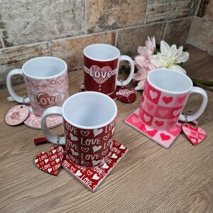 K&ouml;nnte beinhalten: Vier wei&szlig;e Keramikbecher mit verschiedenen roten und rosa "Love"-Designs und passenden Untersetzern. Jeder Becher hat einen wei&szlig;en Griff und einen dekorativen Anh&auml;nger. Die Becher sind auf einer Holzoberfl&auml;che angeordnet.