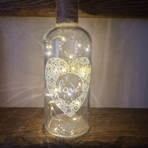 May include: A clear glass bottle with a heart design and the word "love" inside. The bottle is wrapped with twine and contains a string of fairy lights.