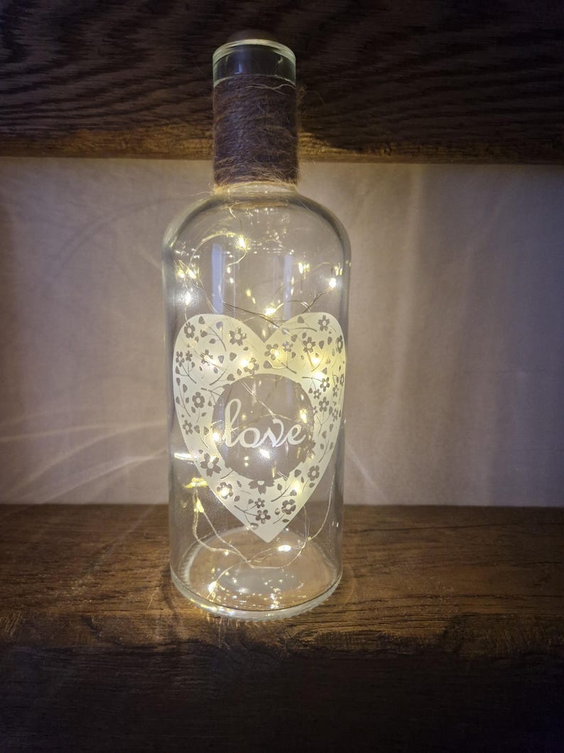 May include: A clear glass bottle with a heart design and the word "love" inside. The bottle is wrapped with twine and contains a string of fairy lights.