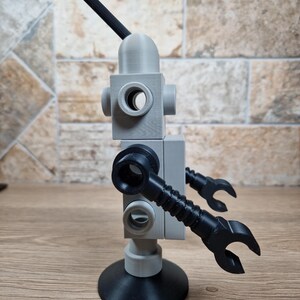 May include: A 3D-printed robot toy in gray and black. The robot has a rounded head with a black antenna, a block-shaped body with circular openings, and articulated arms ending in claw-like hands. It stands on a circular base.