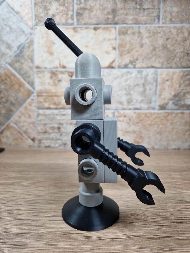 May include: A 3D-printed robot toy in gray and black. The robot has a rounded head with a black antenna, a block-shaped body with circular openings, and articulated arms ending in claw-like hands. It stands on a circular base.