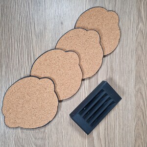 K&ouml;nnte beinhalten: Set aus vier Korkuntersetzern mit schwarzem Rand, in einer Reihe angeordnet. Ein schwarzer Halter befindet sich neben den Untersetzern. Die Untersetzer und der Halter liegen auf einer hellbraunen Holzoberfl&auml;che.