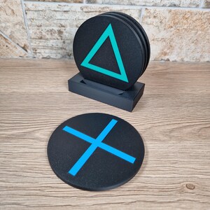 K&ouml;nnte beinhalten: Ein Set aus runden, schwarzen Untersetzern mit leuchtend cyanfarbenen geometrischen Symbolen. Ein Untersetzer zeigt ein Dreieck, ein anderer ein X. Die Untersetzer befinden sich in einem schwarzen rechteckigen Halter, einer liegt flach davor.