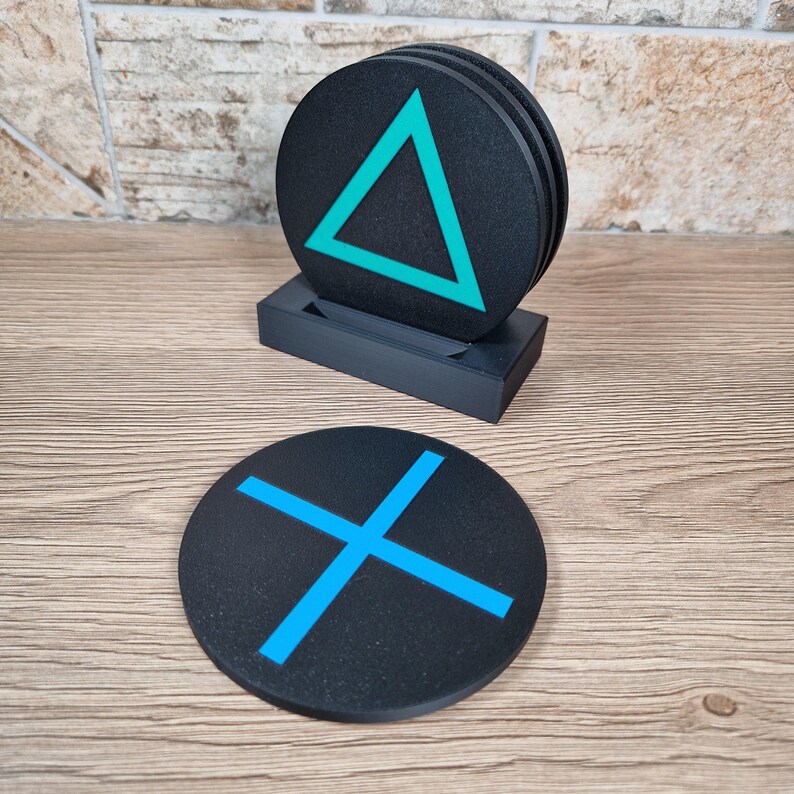 K&ouml;nnte beinhalten: Ein Set aus runden, schwarzen Untersetzern mit leuchtend cyanfarbenen geometrischen Symbolen. Ein Untersetzer zeigt ein Dreieck, ein anderer ein X. Die Untersetzer befinden sich in einem schwarzen rechteckigen Halter, einer liegt flach davor.