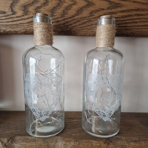May include: Two clear glass bottles with a white heart design and the word "love" etched on them. The bottles are wrapped with twine and have a string of lights inside.