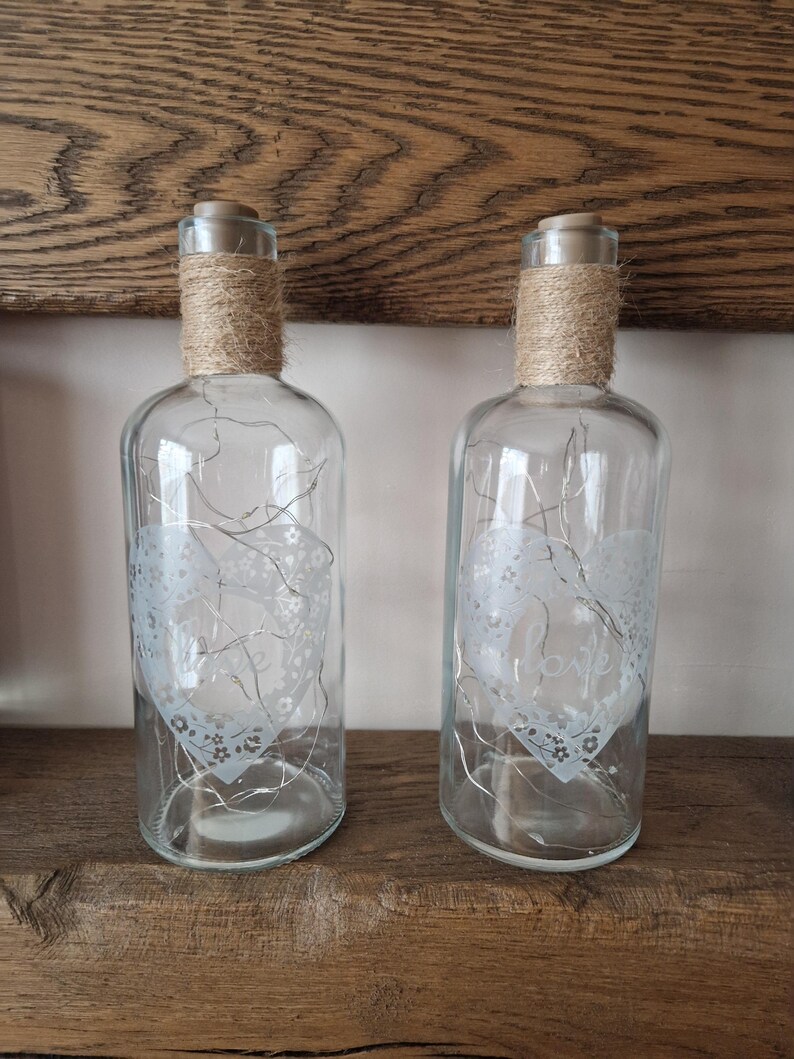 May include: Two clear glass bottles with a white heart design and the word "love" etched on them. The bottles are wrapped with twine and have a string of lights inside.