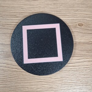 K&ouml;nnte beinhalten: Ein kreisf&ouml;rmiger schwarzer Untersetzer mit strukturierter Oberfl&auml;che und einem zentrierten hellrosa quadratischen Umriss. Das Design ist schlicht und geometrisch und bietet eine flache Oberfl&auml;che f&uuml;r Getr&auml;nke. Der Untersetzer hat ein mattes Finish.