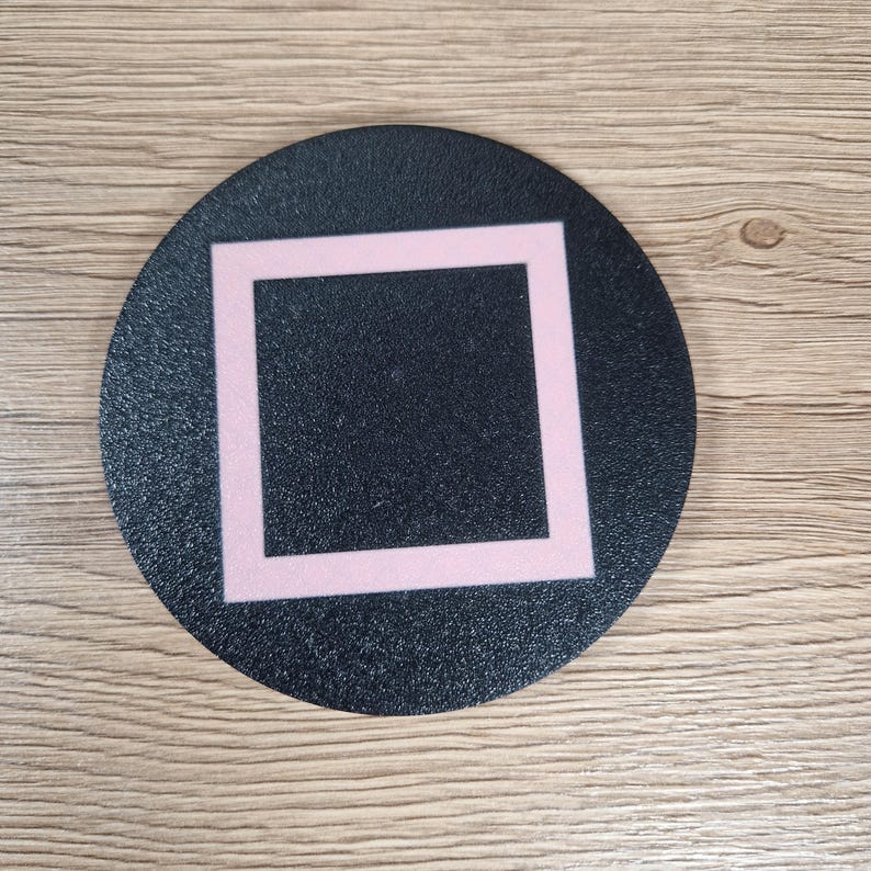 K&ouml;nnte beinhalten: Ein kreisf&ouml;rmiger schwarzer Untersetzer mit strukturierter Oberfl&auml;che und einem zentrierten hellrosa quadratischen Umriss. Das Design ist schlicht und geometrisch und bietet eine flache Oberfl&auml;che f&uuml;r Getr&auml;nke. Der Untersetzer hat ein mattes Finish.