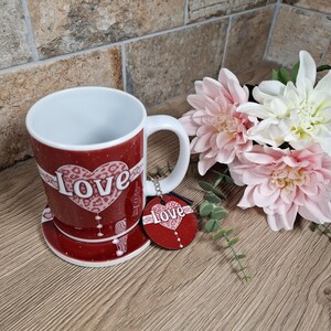May include: A red ceramic mug with a white handle and a matching coaster. The mug and coaster feature a white heart with the word "love" and a leopard print design. A red heart-shaped keychain with the word "love" hangs next to the mug.
