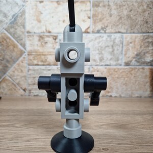 May include: A gray and black 3D-printed robot toy with a circular base and a long antenna. The robot has a cylindrical body with circular and rectangular features. The toy is made of plastic and sits on a wooden surface.