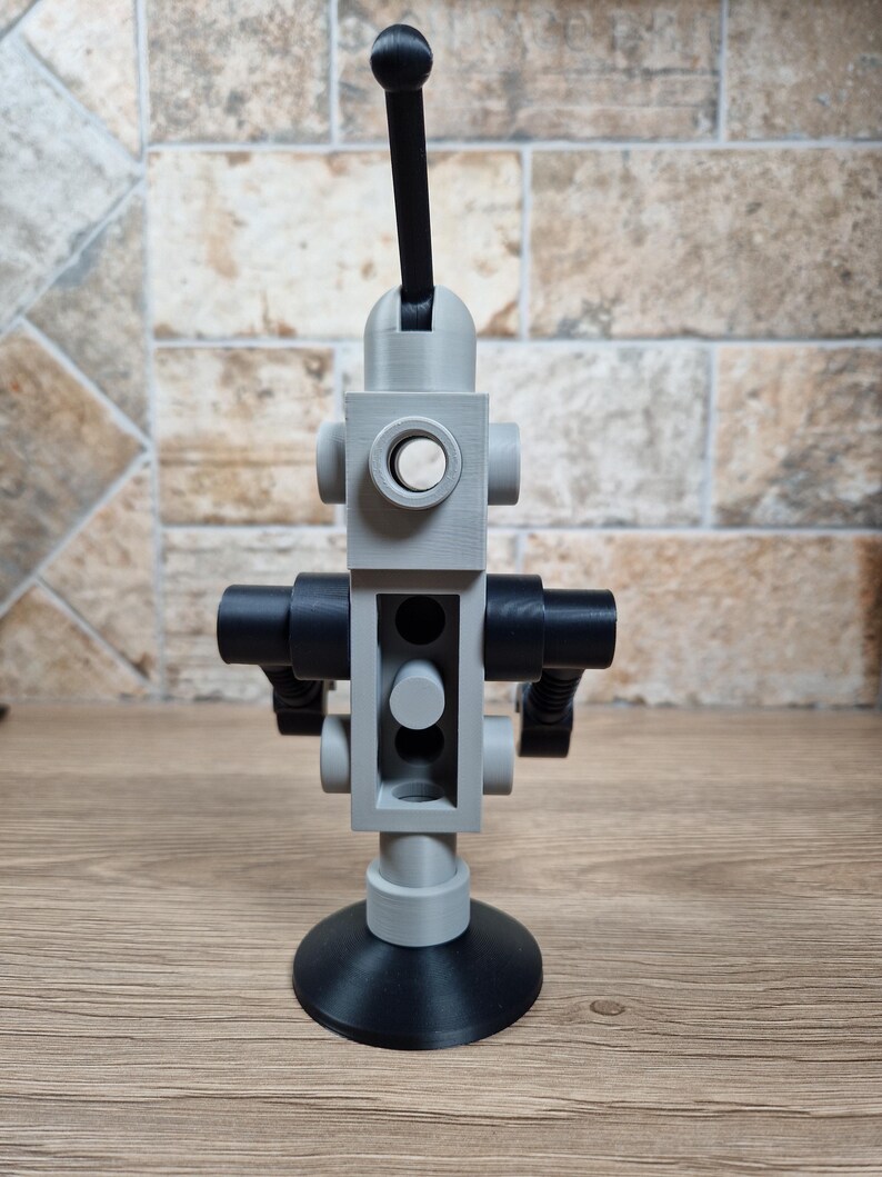 May include: A gray and black 3D-printed robot toy with a circular base and a long antenna. The robot has a cylindrical body with circular and rectangular features. The toy is made of plastic and sits on a wooden surface.