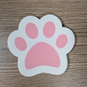 Op de afbeelding: Een wit en roze pootafdruk-vormige onderzetter. De pootafdruk heeft een witte omtrek en roze kussentjes. De onderzetter ligt op een houten oppervlak.