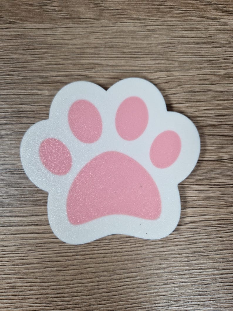 Op de afbeelding: Een wit en roze pootafdruk-vormige onderzetter. De pootafdruk heeft een witte omtrek en roze kussentjes. De onderzetter ligt op een houten oppervlak.