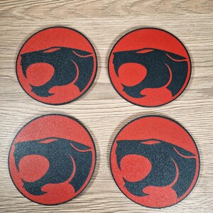 May include: Four round coasters with a red background and a black panther head design. The coasters are arranged in a square pattern and are made of a textured material. The design is a stylized illustration of a panther's head.