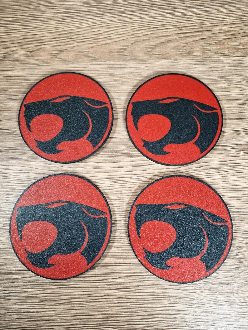 May include: Four round coasters with a red background and a black panther head design. The coasters are arranged in a square pattern and are made of a textured material. The design is a stylized illustration of a panther's head.