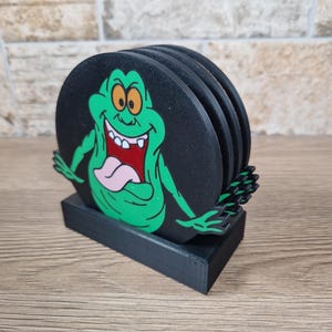 May include: Set of four black coasters featuring a green cartoon ghost character with orange eyes, a pink tongue, and a wide grin. The coasters are round and rest on a black rectangular base.