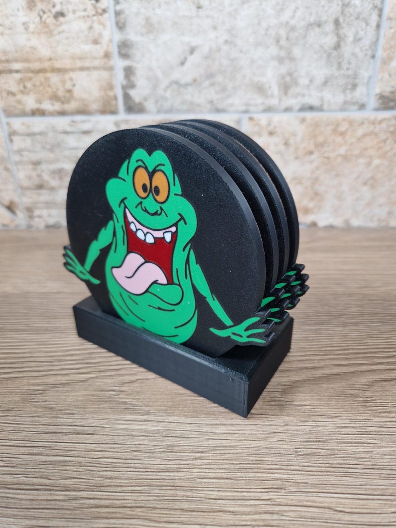 May include: Set of four black coasters featuring a green cartoon ghost character with orange eyes, a pink tongue, and a wide grin. The coasters are round and rest on a black rectangular base.