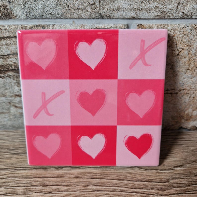 May include: A square, pink and red decorative tile with a tic-tac-toe design. The squares alternate between light pink and red, with hearts and X's in each square. The tile is likely ceramic or a similar material.