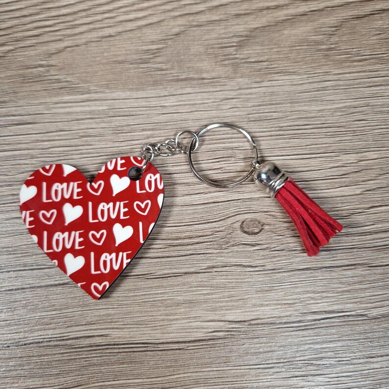 K&ouml;nnte beinhalten: Ein herzf&ouml;rmiger Schl&uuml;sselanh&auml;nger mit rotem Hintergrund und wei&szlig;em "LOVE"-Text und Herzgrafiken. Der Schl&uuml;sselanh&auml;nger hat einen silbernen Ring, eine Kette und eine rote Quaste. Der Schl&uuml;sselanh&auml;nger liegt auf einer Holzoberfl&auml;che.
