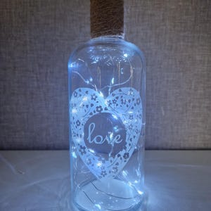 May include: A clear glass bottle with a white heart design and the word "love" inside. The bottle is wrapped with twine and contains white fairy lights.