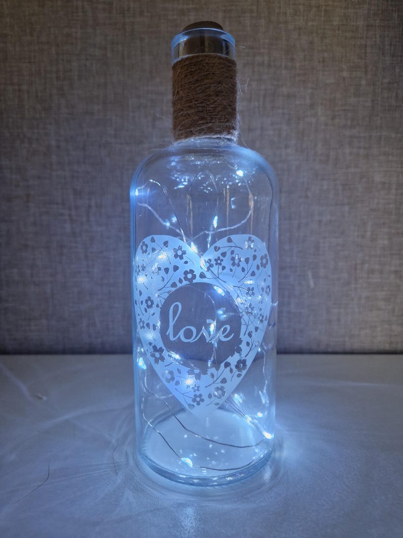 May include: A clear glass bottle with a white heart design and the word "love" inside. The bottle is wrapped with twine and contains white fairy lights.