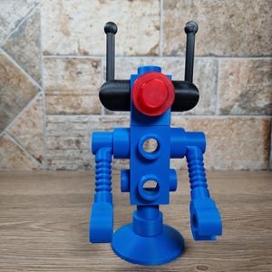 May include: A blue robot toy with a red circular head and black antennae. The robot has a round base and articulated arms. The toy is made of plastic and is designed to resemble a retro robot.