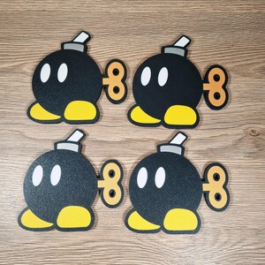May include: Four black cartoon bomb figures with white oval eyes, yellow feet, and a gray fuse. Each bomb has a brown, figure-eight-shaped winder on its side. The bombs are arranged on a light brown wooden surface.