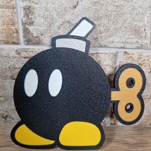 May include: A black cartoon bomb with white eyes, yellow feet, and a silver and white fuse. The bomb has a gold wind-up key on its side. The design is reminiscent of a video game character.