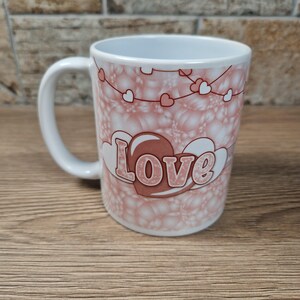May include: White ceramic mug with a pink and white heart design. The word "Love" is displayed in a decorative font on the front of the mug. The mug has a white handle and a light pink interior.