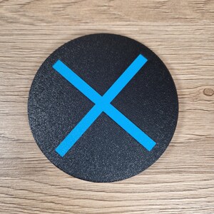K&ouml;nnte beinhalten: Ein kreisf&ouml;rmiger, schwarzer Untersetzer mit strukturierter Oberfl&auml;che und einem leuchtend blauen X-Symbol in der Mitte. Der Untersetzer hat eine flache, runde Form mit sauberen Kanten und einer matten Oberfl&auml;che f&uuml;r ein schlichtes, funktionales Design.
