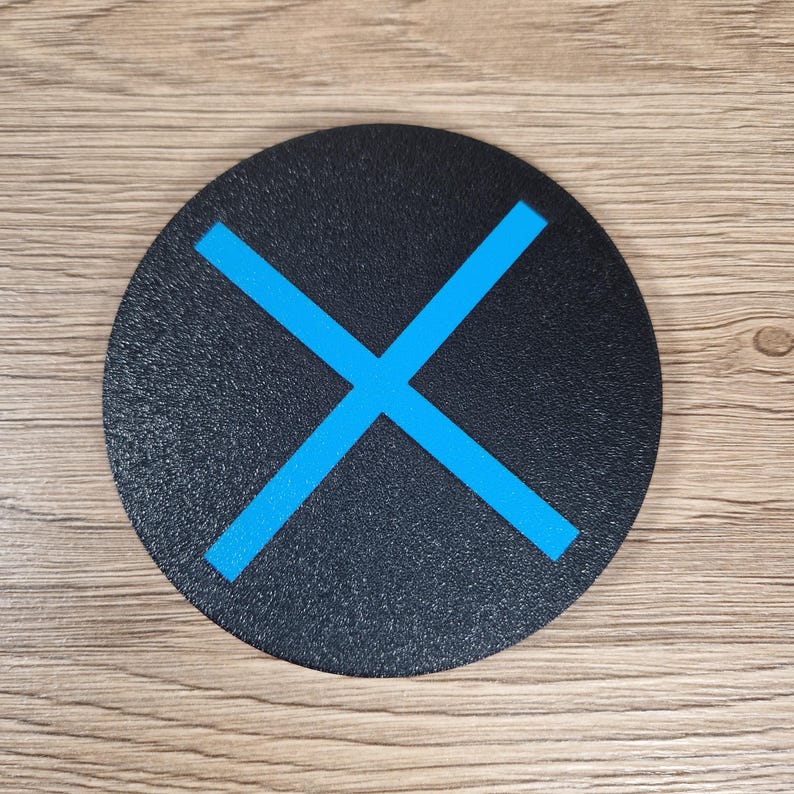 K&ouml;nnte beinhalten: Ein kreisf&ouml;rmiger, schwarzer Untersetzer mit strukturierter Oberfl&auml;che und einem leuchtend blauen X-Symbol in der Mitte. Der Untersetzer hat eine flache, runde Form mit sauberen Kanten und einer matten Oberfl&auml;che f&uuml;r ein schlichtes, funktionales Design.