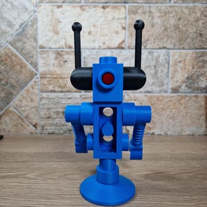 May include: A blue robot figurine with a red eye and black antennae. The robot has a cylindrical body with two arms and a circular base. The robot is made of plastic and sits on a wooden surface.