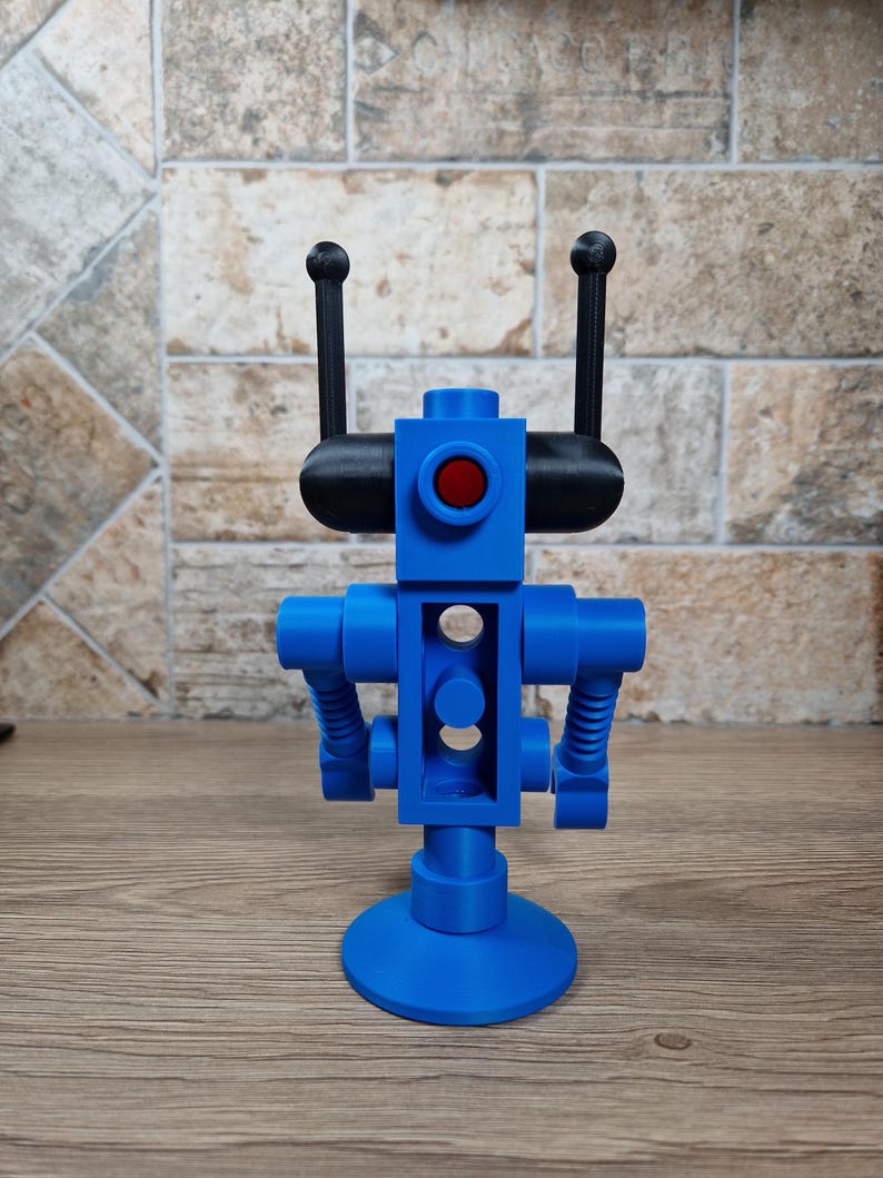May include: A blue robot figurine with a red eye and black antennae. The robot has a cylindrical body with two arms and a circular base. The robot is made of plastic and sits on a wooden surface.