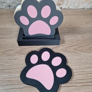 Op de afbeelding: Een set pootvormige onderzetters in een houder. De onderzetters zijn zwart met roze voetzolen. De houder is zwart. De onderzetters zijn in een houder gestapeld, met &eacute;&eacute;n onderzetter plat op het oppervlak.