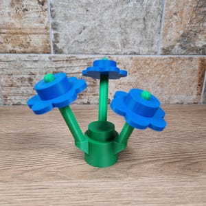 May include: A 3D-printed flower sculpture featuring three blue flower heads with green centers and stems. The base is green, and the sculpture sits on a wooden surface. The flowers have a unique, stylized design.