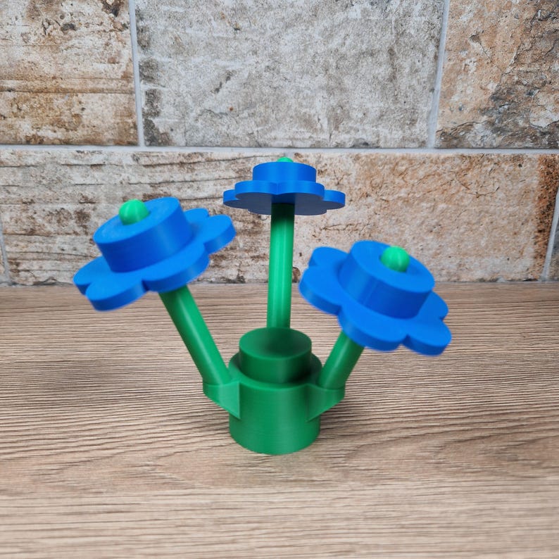 May include: A 3D-printed flower sculpture featuring three blue flower heads with green centers and stems. The base is green, and the sculpture sits on a wooden surface. The flowers have a unique, stylized design.