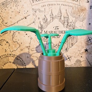 May include: A 3D-printed, green and brown decorative item. The item features a barrel-shaped base with a green, stylized plant design on top. The background includes a map with text that reads "Wormtail, Padfoot & Prongs".