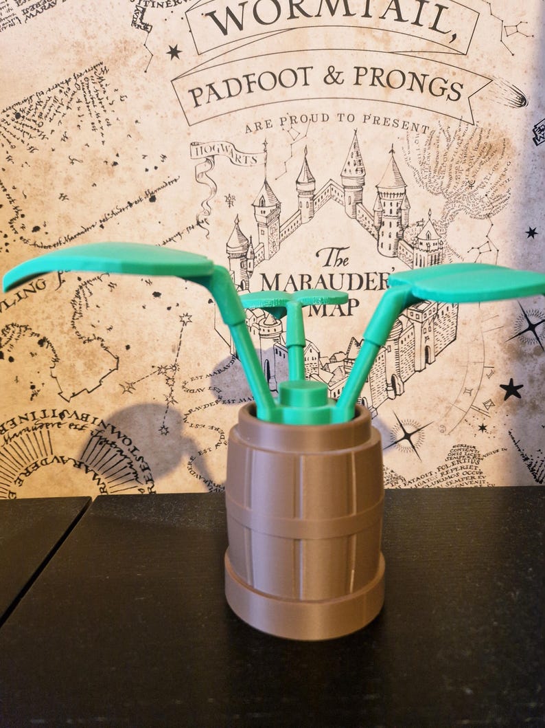 May include: A 3D-printed, green and brown decorative item. The item features a barrel-shaped base with a green, stylized plant design on top. The background includes a map with text that reads "Wormtail, Padfoot & Prongs".