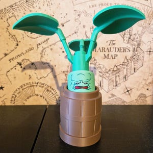 May include: A 3D-printed Mandrake plant figure, featuring a green plant with large leaves and a face, housed in a brown barrel-shaped pot. The background includes a map with text that reads "PADFOOT & PRONGS" and "The Marauders Map."
